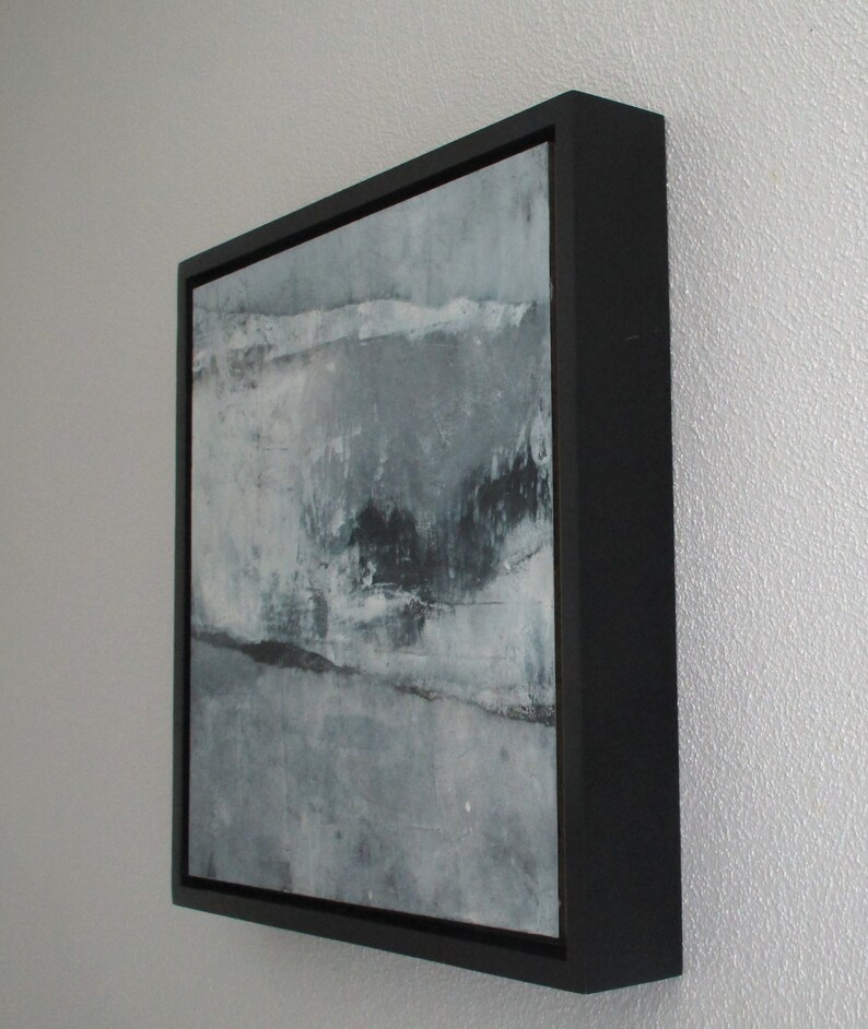 Op de afbeelding: Abstract kunstwerk in een zwarte lijst. Het kunstwerk toont een gestructureerd, gelaagd ontwerp in grijstinten, wit en zwart. Het schilderij heeft een moderne esthetiek, met zichtbare penseelstreken en een minimalistische stijl. De lijst is vierkant en heeft een matte afwerking.