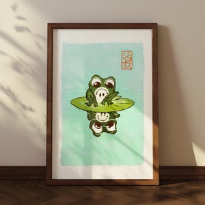 Peut inclure: Impression d'art encadrée représentant deux grenouilles vertes dessinées sur une feuille de nénuphar, sur fond bleu clair. L'œuvre comprend des caractères japonais en haut à droite et est encadrée dans un cadre en bois marron.