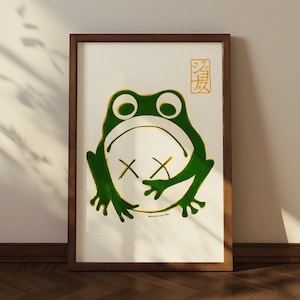 Peut inclure: Impression d'art encadrée représentant une grenouille verte avec des yeux barrés sur fond blanc. L'œuvre est encadrée dans un cadre en bois marron. Du texte japonais est visible en haut à droite.