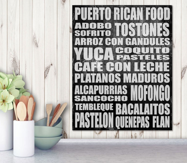 May include: Black rectangular sign with white text listing Puerto Rican food items like 'Mofongo' and 'Pasteles'. The sign is set against a white wooden background, with kitchen utensils and a vase of flowers in the foreground.