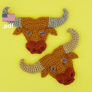 Puede incluir: Dos cabezas de toro de crochet con pelo marrón y naranja, cuernos grises y ojos azules. Las cabezas de toro están sobre un fondo amarillo.