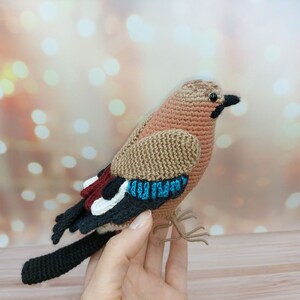 May include: A crocheted stuffed bird with brown, black, white, and blue details. The bird has a long tail and a small beak.