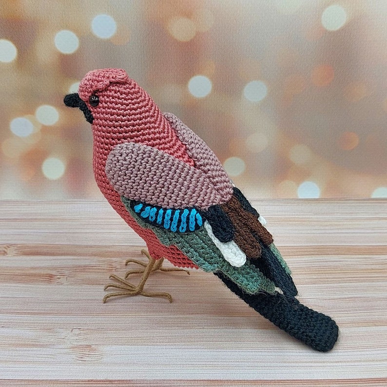 May include: A crocheted bird toy with a pink body, blue and green wings, and a black tail. The bird is standing on a wooden surface.
