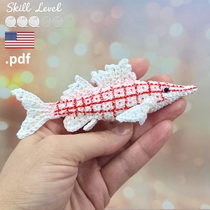 May include: A small, white crocheted fish with a red grid pattern across its body, featuring a pointed snout, a dark eye, and textured fins. The fish is crafted with fine yarn and displays intricate stitching details throughout its entire form.