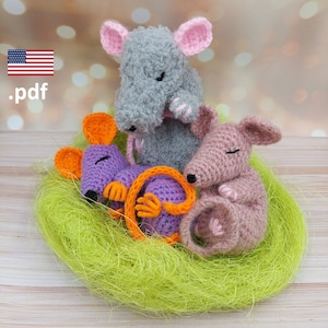 Könnte beinhalten: Drei gehäkelte Mäuse in einem grünen Nest. Die Mäuse sind grau, lila und rosa. Die graue Maus hat rosa Ohren und eine rosa Nase. Die lila Maus hat orange Ohren und einen eingerollten Schwanz. Die rosa Maus hat rosa Ohren und einen eingerollten Schwanz.