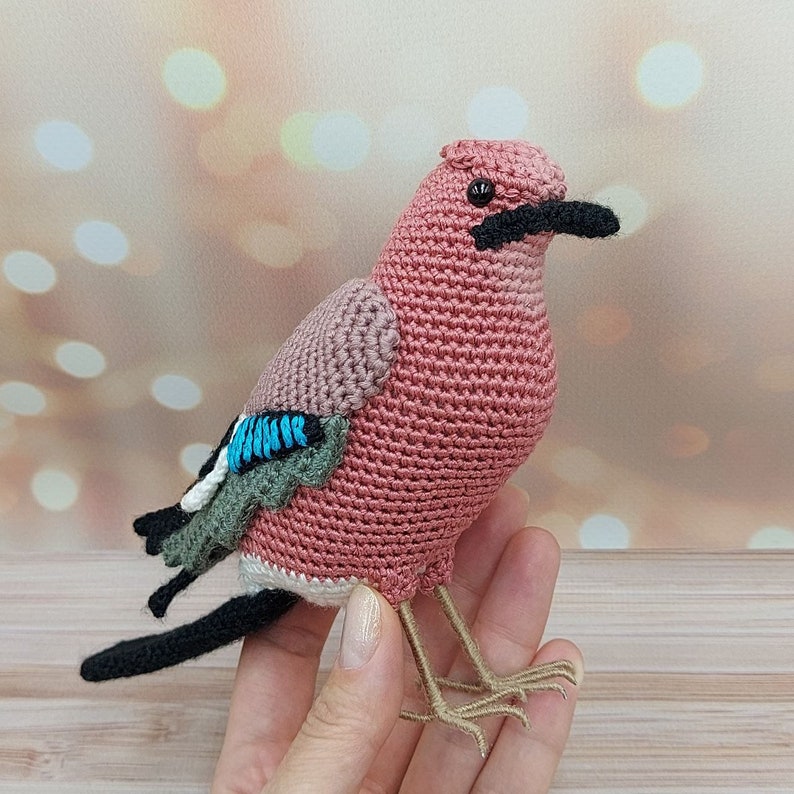 May include: A crocheted bird toy with a pink body, black eyes, and a black beak. The bird has blue and green wings and brown legs.