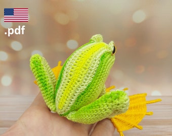 May include: A crocheted green and yellow frog with a yellow crocheted tail. The frog is sitting on a hand.