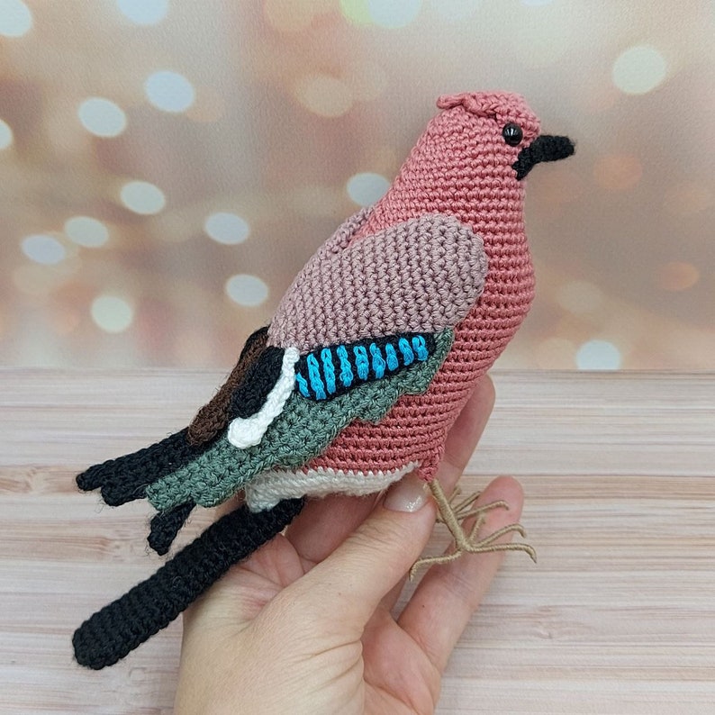 May include: A crocheted amigurumi bird toy with a pink body, brown, green, and blue wings, and black legs.