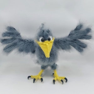 Puede incluir: Un pájaro de crochet gris con pico y patas amarillas. El pájaro tiene grandes ojos negros y está de pie con las alas extendidas.