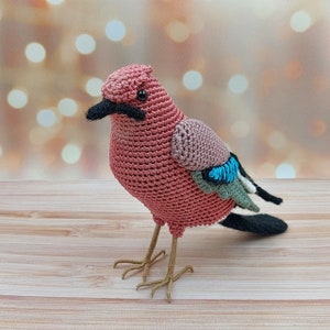 May include: A crocheted bird toy with pink, brown, and blue accents. The bird has a long, black beak and a small, black eye.