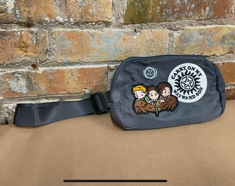 May include: Gray fanny pack with an embroidered patch that says "Carry On My Wayward Son" and features three cartoon characters. The patch is surrounded by a white circle with a black star in the center.