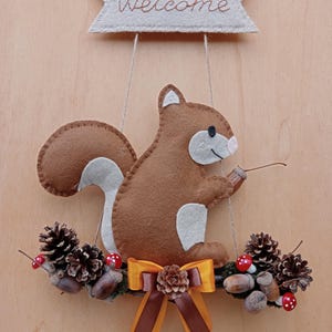 May include: A felt squirrel welcome sign. The brown squirrel has white accents and holds a twig. It sits on a branch with pine cones, acorns, and a decorative bow. The sign has a banner that says 'Welcome' and is suspended by string.