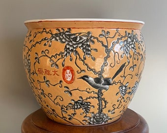 May include: A large, orange ceramic planter with a black and white floral design and a bird perched on a branch. The planter has Chinese characters that read "福祿大" in red.