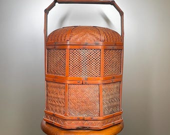May include: A vintage, reddish-brown bamboo basket with a handle. The basket has a domed lid and a rectangular shape with woven and lattice-style panels. It is displayed on a wooden pedestal.