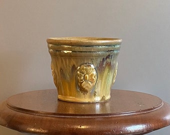 May include: A glazed ceramic planter with a brown and yellow glaze. The planter has a rounded shape and features embossed floral designs.