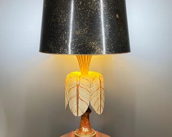 May include: A decorative table lamp with a black lampshade speckled with gold. The lamp features a unique design with a floral-like base in cream and gold, and a wooden stand. The lamp is placed on a small, round wooden table.