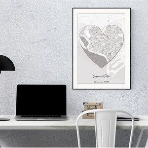 May include: A black and white map of a city in the shape of a heart, framed in black, hanging on a white wall above a white desk with a black lamp and a laptop. The map is titled "Suzanne & Peter" with the text "Our love began... 2022".