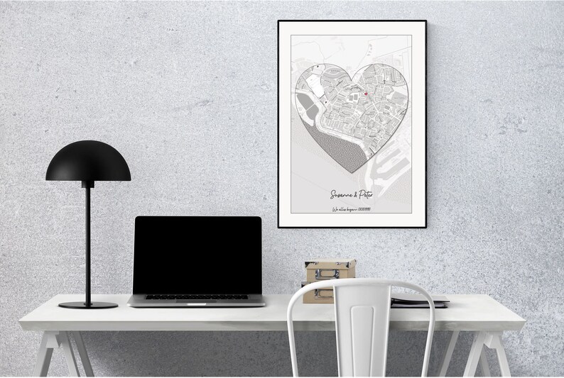 May include: A black and white map of a city in the shape of a heart, framed in black, hanging on a white wall above a white desk with a black lamp and a laptop. The map is titled "Suzanne & Peter" with the text "Our love began... 2022".