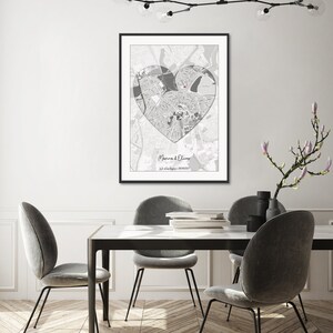 May include: A black and white map of a city in the shape of a heart with the names "Marina & Oliver" and a date printed below. The map is framed and hanging on a white wall above a dining table with four chairs.