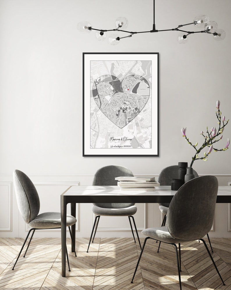 May include: A black and white map of a city in the shape of a heart with the names "Marina & Oliver" and a date printed below. The map is framed and hanging on a white wall above a dining table with four chairs.