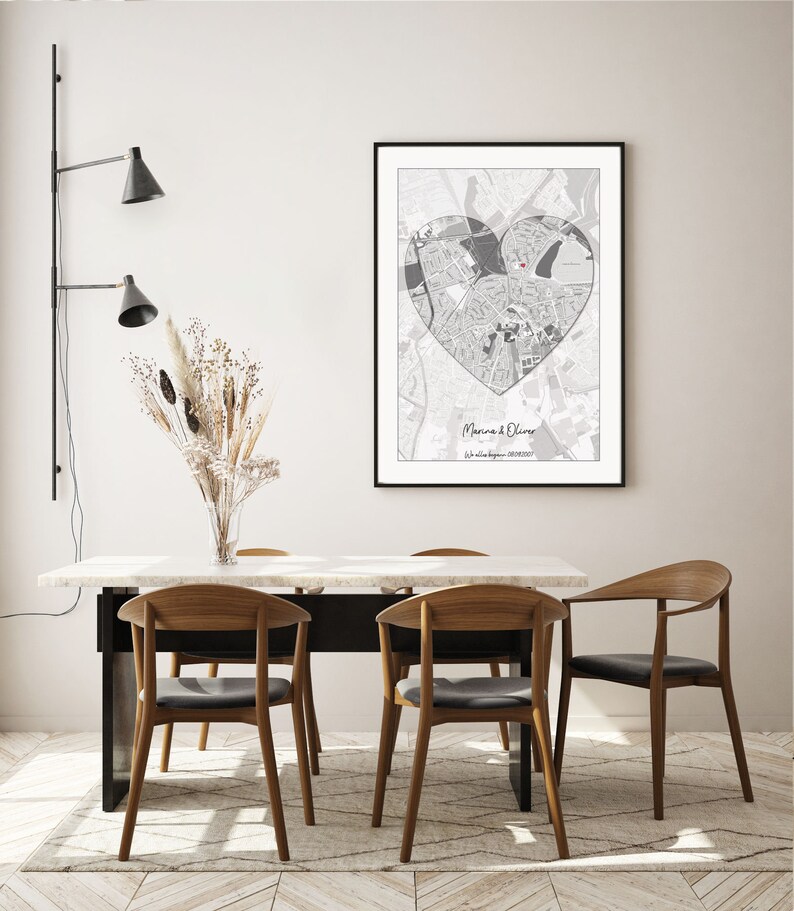 May include: A black and white map of a city in the shape of a heart with a red dot in the center. The map is framed and hanging on a white wall above a dining table with four chairs.
