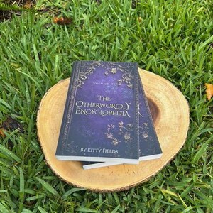 May include: Two books titled "The Otherworldly Encyclopedia" by Kitty Fields, Volume Def, with a purple cover and gold floral accents, rest on a round wooden slice. The books are set against a backdrop of green grass.