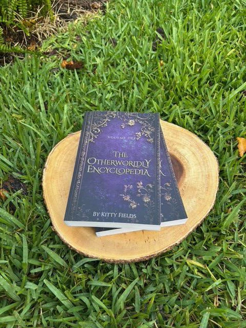May include: Two books titled "The Otherworldly Encyclopedia" by Kitty Fields, Volume Def, with a purple cover and gold floral accents, rest on a round wooden slice. The books are set against a backdrop of green grass.
