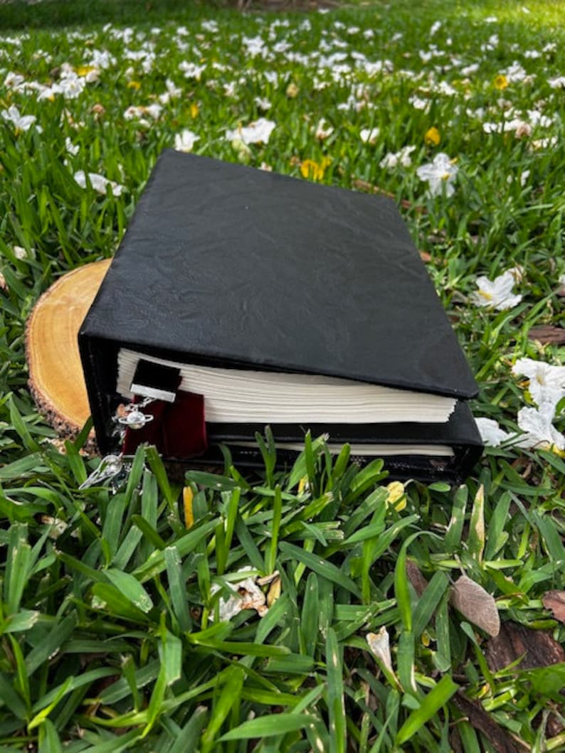 Pu&ograve; includere: Un diario rilegato in pelle nera con un segnalibro in nastro bordeaux. Il diario &egrave; appoggiato su un disco di legno in un letto di erba verde e petali di fiori bianchi.