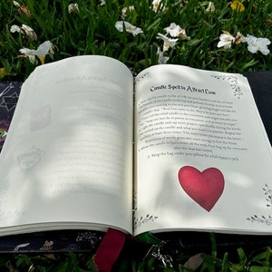 Pu&ograve; includere: Libro aperto con un'illustrazione di cuore rosso sulla pagina. Il libro &egrave; intitolato "Candle Spell to Attract Love" e contiene istruzioni per un incantesimo con la candela. Il libro &egrave; appoggiato su un letto di erba verde e fiori bianchi.