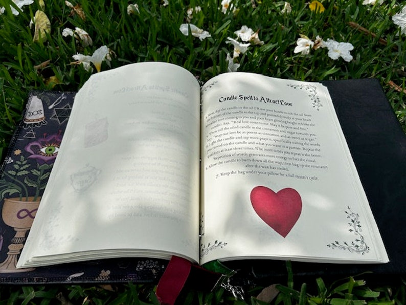 Pu&ograve; includere: Libro aperto con un'illustrazione di cuore rosso sulla pagina. Il libro &egrave; intitolato "Candle Spell to Attract Love" e contiene istruzioni per un incantesimo con la candela. Il libro &egrave; appoggiato su un letto di erba verde e fiori bianchi.