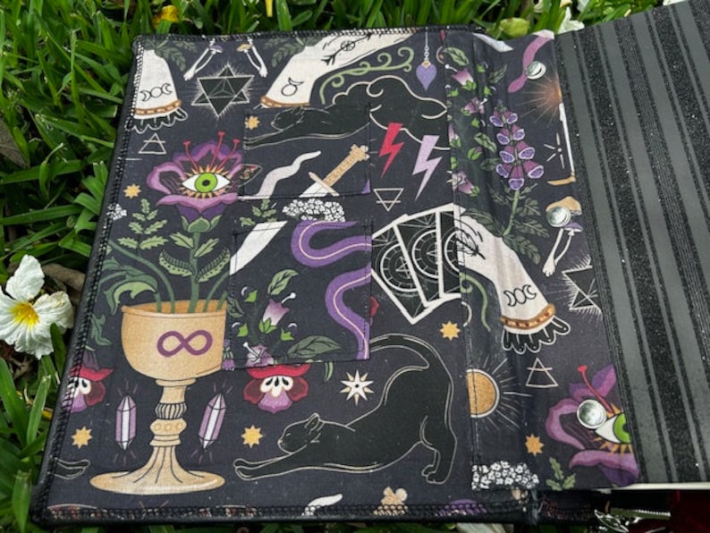 Pu&ograve; includere: Una copertina per diario in tessuto nero con una tasca sul davanti. La copertina presenta un motivo ripetitivo di vari simboli, tra cui un gatto, un calice, un serpente, un pugnale e una mano con un occhio. Lo sfondo &egrave; nero con accenti viola e dorati.