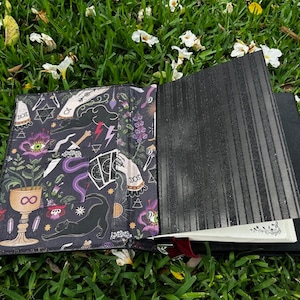 Pu&ograve; includere: Un diario nero con una copertina a motivi che presenta un gatto nero, un calice, un occhio e altri simboli occulti. La copertina &egrave; aperta per rivelare una pagina bianca.