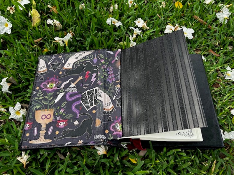 Pu&ograve; includere: Un diario nero con una copertina a motivi che presenta un gatto nero, un calice, un occhio e altri simboli occulti. La copertina &egrave; aperta per rivelare una pagina bianca.