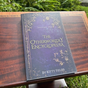 May include: A hardcover book titled "The Otherworldly Encyclopedia" by Kitty Fields. The book has a dark purple cover with gold floral designs and text. The book is open on a dark wood table.