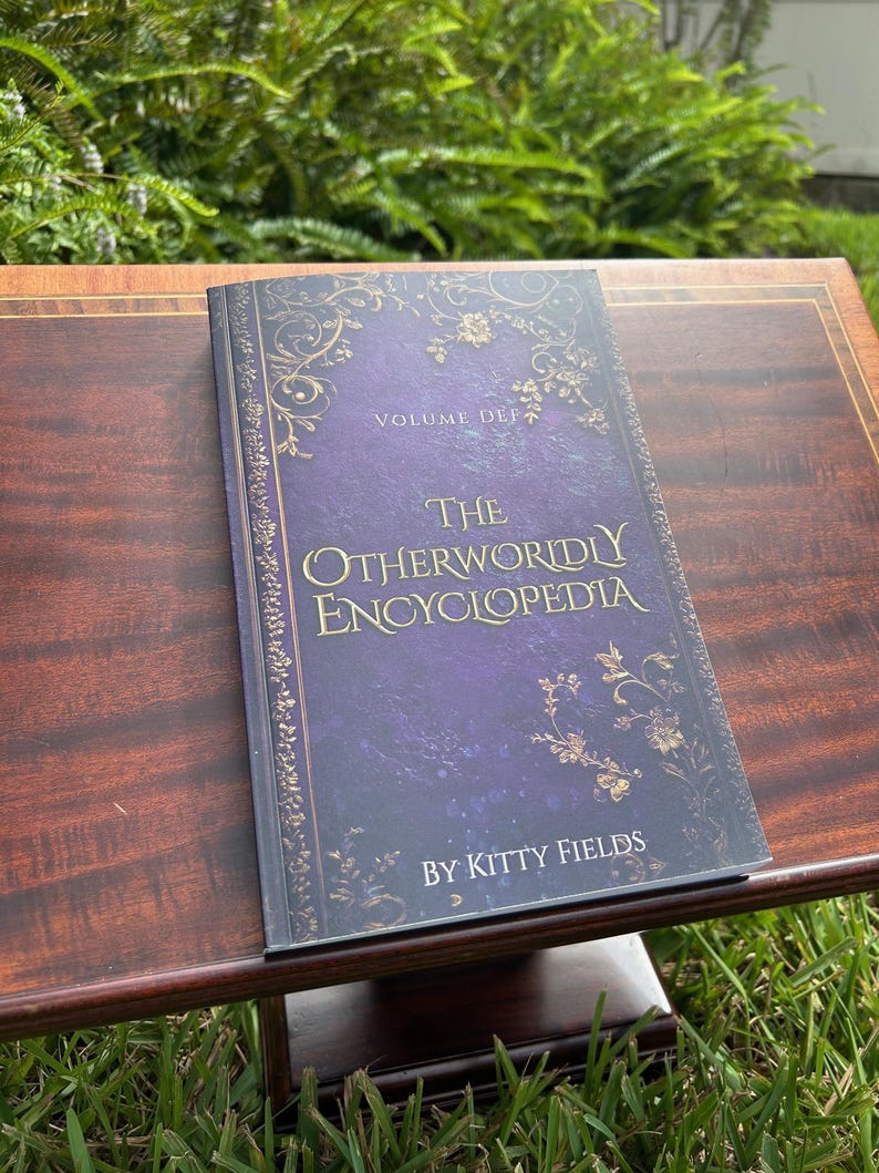 May include: A hardcover book titled "The Otherworldly Encyclopedia" by Kitty Fields. The book has a dark purple cover with gold floral designs and text. The book is open on a dark wood table.