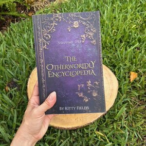 May include: A hardcover book titled "The Otherworldly Encyclopedia" with a purple and gold cover. The book is held on a wooden slice against a backdrop of green grass. The book's cover features ornate floral designs and the author's name, Kitty Fields.