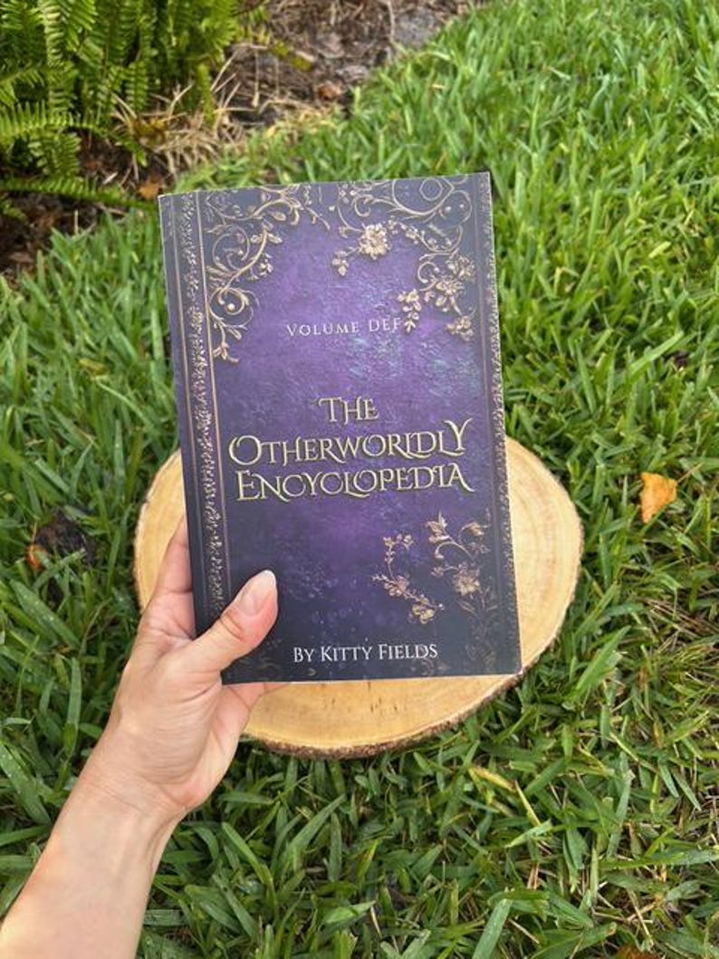 May include: A hardcover book titled "The Otherworldly Encyclopedia" with a purple and gold cover. The book is held on a wooden slice against a backdrop of green grass. The book's cover features ornate floral designs and the author's name, Kitty Fields.