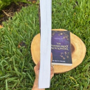 May include: A hardcover book with white-edged pages is held in front of a book with a purple cover. The book on the bottom has gold lettering and the title "The Otherworldly Encyclopedia" by Kitty Fields.