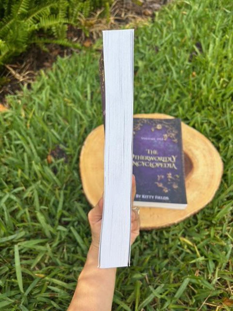 May include: A hardcover book with white-edged pages is held in front of a book with a purple cover. The book on the bottom has gold lettering and the title "The Otherworldly Encyclopedia" by Kitty Fields.