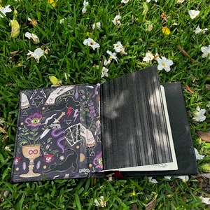 Pu&ograve; includere: Un diario in pelle nera con una copertina illustrata colorata che presenta un gatto nero, una luna crescente, un simbolo di infinito e altri simboli mistici. Il diario &egrave; aperto e appoggiato su un letto di erba verde con fiori bianchi.