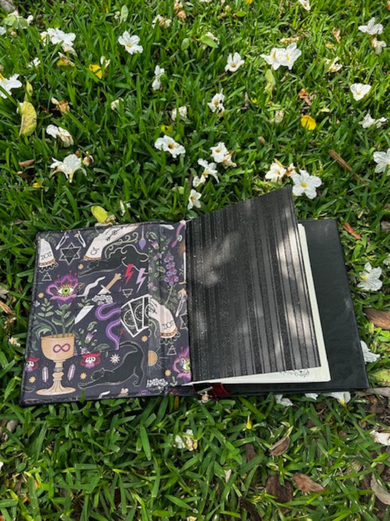 Pu&ograve; includere: Un diario in pelle nera con una copertina illustrata colorata che presenta un gatto nero, una luna crescente, un simbolo di infinito e altri simboli mistici. Il diario &egrave; aperto e appoggiato su un letto di erba verde con fiori bianchi.