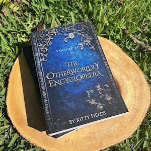 May include: A blue hardcover book with gold lettering titled "The Otherworldly Encyclopedia, Volume BC" by Kitty Fields. The book is sitting on a wooden slice.