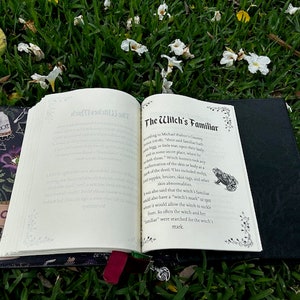 Pu&ograve; includere: Un libro aperto con una copertina nera e un interno bianco. Il libro &egrave; sdraiato su un letto di erba verde con fiori bianchi. Il titolo del libro &egrave; "The Witch's Familiar".
