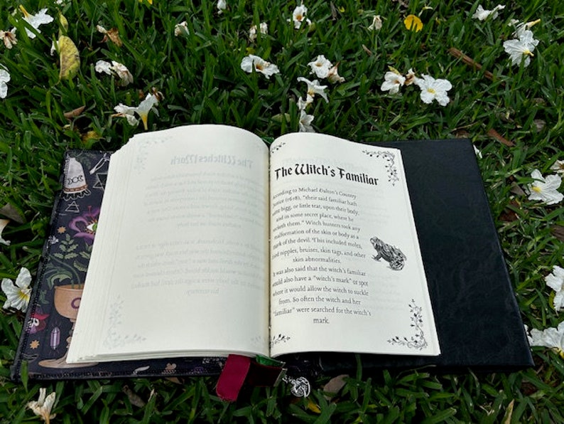 Pu&ograve; includere: Un libro aperto con una copertina nera e un interno bianco. Il libro &egrave; sdraiato su un letto di erba verde con fiori bianchi. Il titolo del libro &egrave; "The Witch's Familiar".