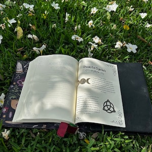 Pu&ograve; includere: Un libro aperto con una copertina nera e un disegno floreale sulla copertina. Il libro &egrave; aperto su un letto di erba verde con fiori bianchi sparsi intorno. Le pagine sono aperte a una sezione intitolata "Simboli Potenti" e mostrano un disegno di un simbolo di luna tripla e un triskel.