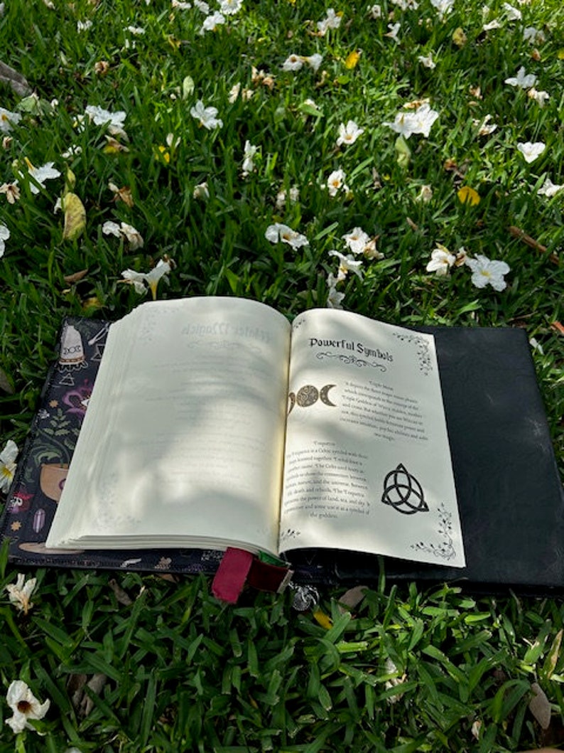 Pu&ograve; includere: Un libro aperto con una copertina nera e un disegno floreale sulla copertina. Il libro &egrave; aperto su un letto di erba verde con fiori bianchi sparsi intorno. Le pagine sono aperte a una sezione intitolata "Simboli Potenti" e mostrano un disegno di un simbolo di luna tripla e un triskel.