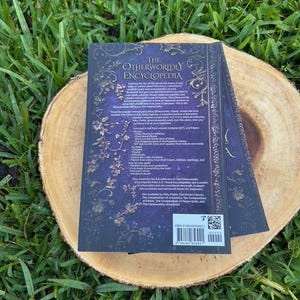 May include: A hardcover book titled "The Otherworldly Encyclopedia" with a dark blue cover and gold floral accents, resting on a wooden slice. The book's title is in white, and the back cover includes text and a barcode.