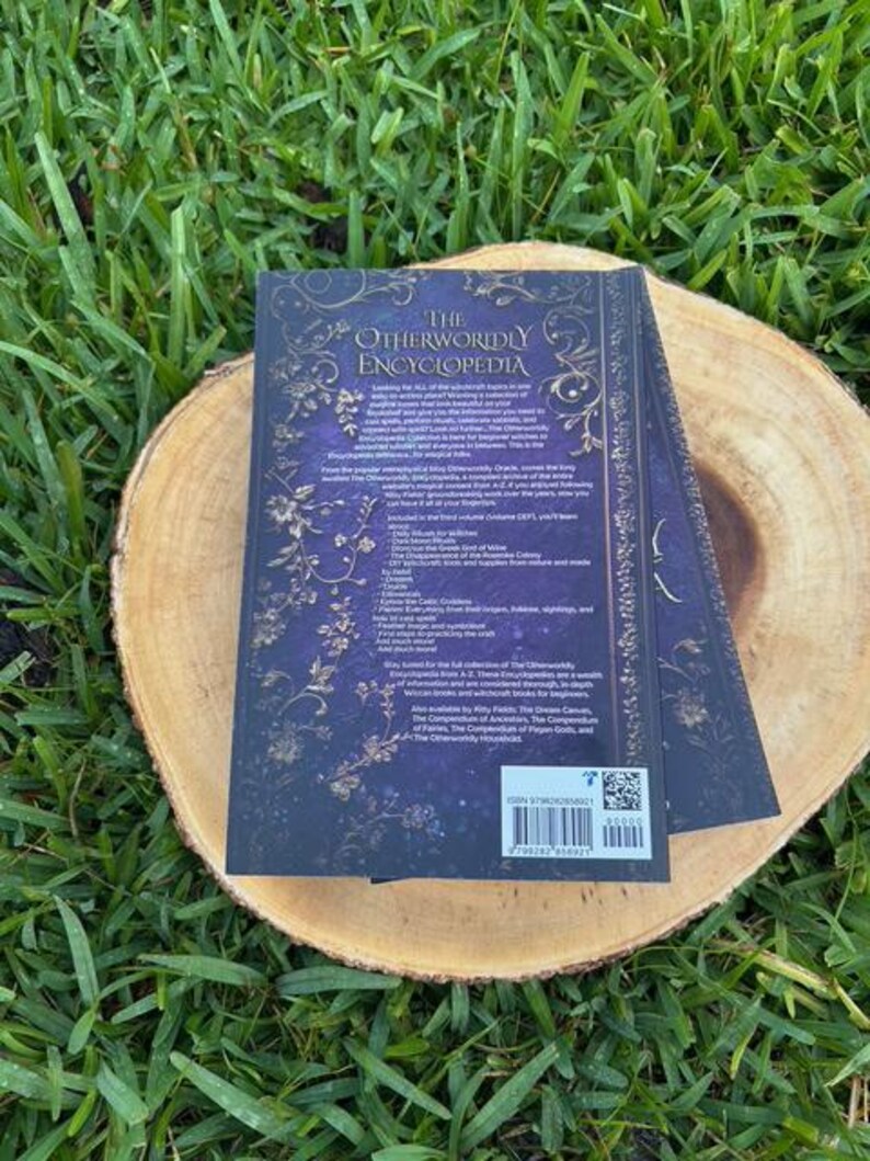 May include: A hardcover book titled "The Otherworldly Encyclopedia" with a dark blue cover and gold floral accents, resting on a wooden slice. The book's title is in white, and the back cover includes text and a barcode.