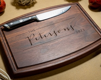 May include: A rectangular dark brown wooden chopping board with a juice groove along the perimeter. The centre is engraved with the text "THE PETERSONS" in a script font, flanked by horizontal lines and the year "2023". A metal knife rests on top.