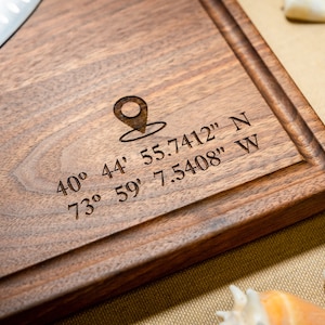 May include: A brown wooden cutting board with a map pin icon and coordinates engraved: 40° 44' 55.7412" N 73° 59' 7.5408" W.